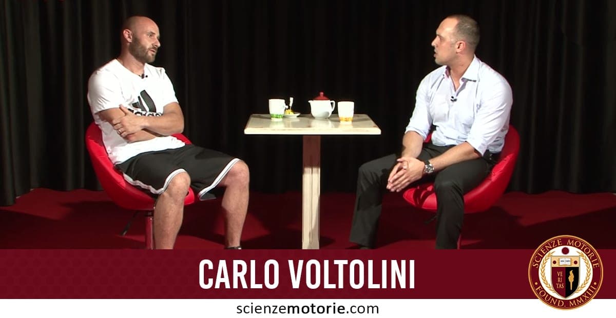 Carlo Voltolini e Giacomo Catalani seduti su poltrone rosse, con un tavolino tra loro con una teiera e una tazza. Sullo sfondo tende scure e in basso il logo di scienzemotorie.com.