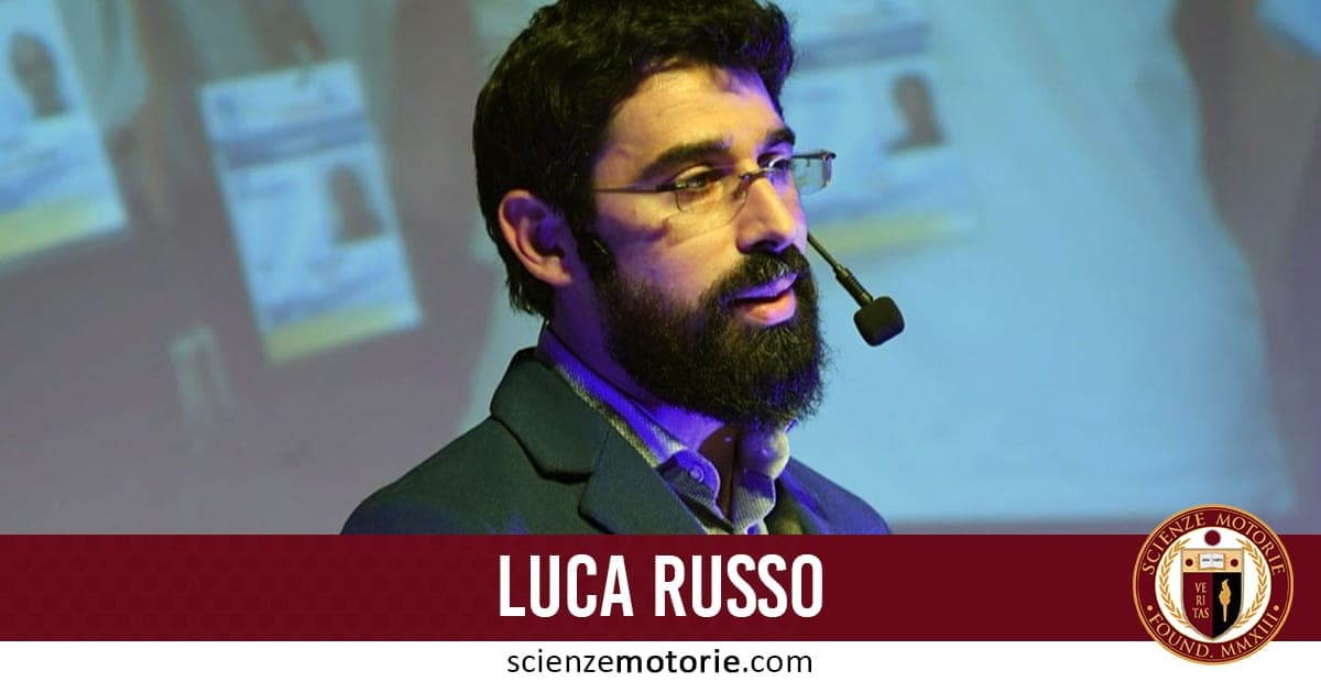Luca Russo durante una presentazione con microfono ad archetto in una sala conferenze, sfondo con proiezione sfocata, logo scienzemotorie.com in basso a destra.